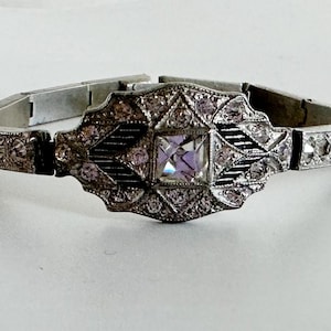 Puede incluir: Un brazalete de estilo Art Déco con una piedra central iridiscente de corte cuadrado. El brazalete de metal plateado presenta un diseño geométrico con pequeñas piedras claras de acento. El brazalete se muestra sobre una superficie blanca.