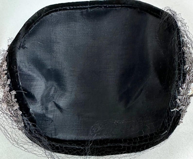 May include: A black hat with a rounded crown and a black veil. The hat is trimmed with small, silver-colored beads. The interior of the hat is lined with a black fabric.