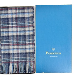 Pode incluir: Um cachecol xadrez azul, cinza e vermelho com franjas, apresentado em uma caixa azul. A caixa apresenta o logotipo da Pendleton e o texto "Always virgin wool".