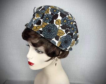 May include: A white beret with a floral pattern in shades of blue, yellow, and brown. The beret has a black ribbon tied around it.