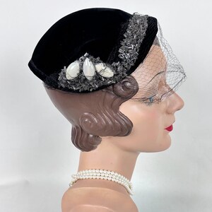 May include: A black velvet hat with a decorative beaded trim and a delicate veil. The hat features white, oval-shaped embellishments and is displayed on a mannequin head. A pearl necklace is also visible.