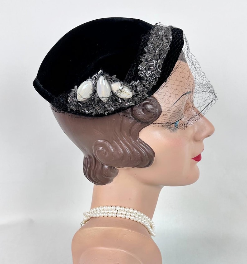 May include: A black velvet hat with a decorative beaded trim and a delicate veil. The hat features white, oval-shaped embellishments and is displayed on a mannequin head. A pearl necklace is also visible.