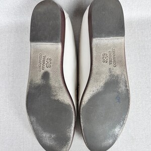 May include: Pair of white leather flats with dark grey soles. The soles show wear and the brand name "SAS TRIPAD COMFORT" is printed on the sole. The shoes have a low heel and a rounded toe.