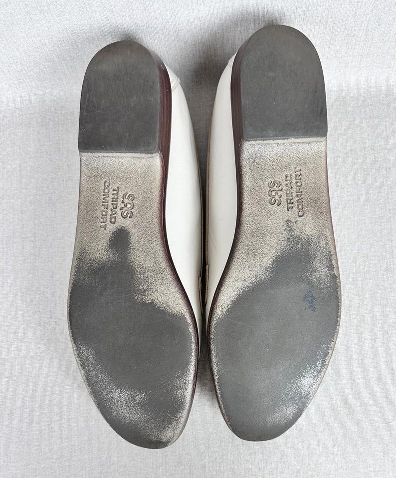 May include: Pair of white leather flats with dark grey soles. The soles show wear and the brand name "SAS TRIPAD COMFORT" is printed on the sole. The shoes have a low heel and a rounded toe.