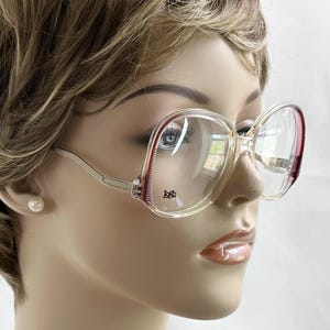 May include: Clear oversized eyeglasses with a light brown frame and red accents. The glasses have a rectangular shape and a silver-colored arm. The logo is visible on the left lens.