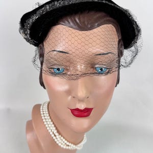 May include: A black velvet hat with a decorative trim and a delicate black netting veil. The hat is displayed on a mannequin head with blue eyes, red lips, and a pearl necklace. A vintage fashion accessory.