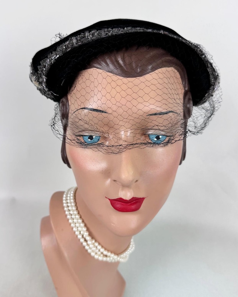 May include: A black velvet hat with a decorative trim and a delicate black netting veil. The hat is displayed on a mannequin head with blue eyes, red lips, and a pearl necklace. A vintage fashion accessory.