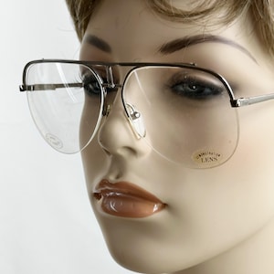 May include: Silver-framed eyeglasses with clear lenses. The glasses have a double bridge design and a rectangular shape. The lenses have a sticker that reads "DEMONSTRATION LENS". The glasses are on a mannequin.