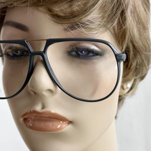 May include: Close-up of a mannequin wearing large, black-rimmed eyeglasses. The glasses have a classic aviator shape with a thin gold bar across the top. The mannequin has short, light brown hair and full, glossy lips.