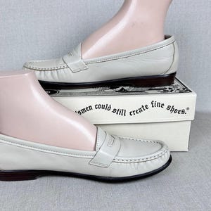 May include: Off-white leather loafers with dark brown soles. The shoes feature a classic penny loafer design with a strap across the vamp. The shoes are displayed on a white box with text that reads "...men could still create fine shoes."
