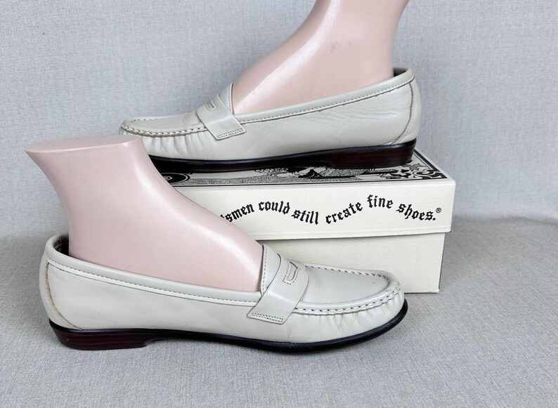May include: Off-white leather loafers with dark brown soles. The shoes feature a classic penny loafer design with a strap across the vamp. The shoes are displayed on a white box with text that reads "...men could still create fine shoes."