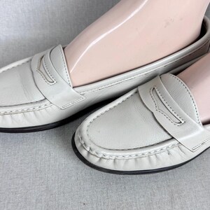 May include: A pair of light grey leather loafers with a classic penny loafer design. The shoes have a stitched trim and a black sole. The shoes are displayed on a light grey surface.