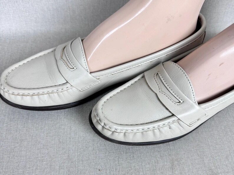 May include: A pair of light grey leather loafers with a classic penny loafer design. The shoes have a stitched trim and a black sole. The shoes are displayed on a light grey surface.