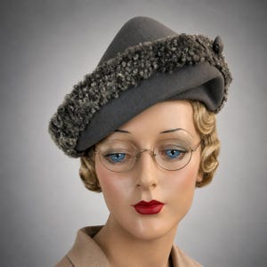 May include: A charcoal gray felt hat featuring a structured, asymmetrical crown and a wide, textured band of dark gray curly faux fur trim. The hat is displayed on a mannequin head wearing thin, gold-rimmed oval eyeglasses with clear lenses.