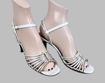 Puede incluir: Sandalias de tiras blancas y doradas con tacón pequeño. Las sandalias tienen una fina correa en el tobillo y múltiples tiras en la parte superior del pie. Los tacones son negros. Las sandalias se exhiben en un pie de maniquí.