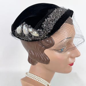 May include: A black velvet hat with a decorative beaded band and three iridescent embellishments. A fine black netting veil drapes over the face. The hat is displayed on a mannequin head with a pearl necklace.
