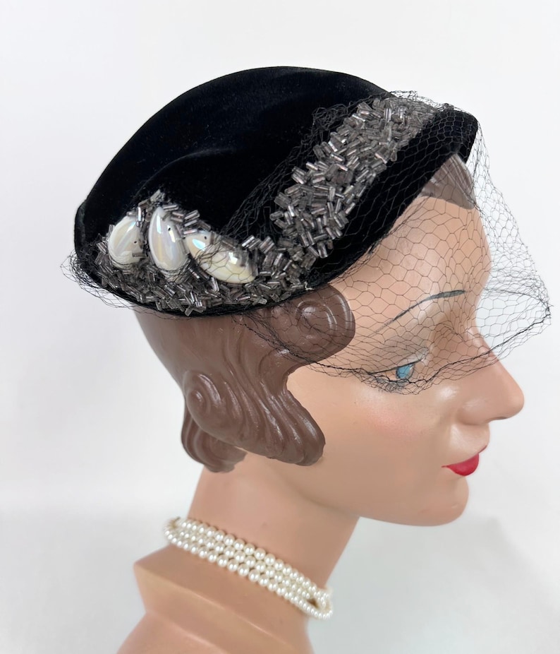 May include: A black velvet hat with a decorative beaded band and three iridescent embellishments. A fine black netting veil drapes over the face. The hat is displayed on a mannequin head with a pearl necklace.