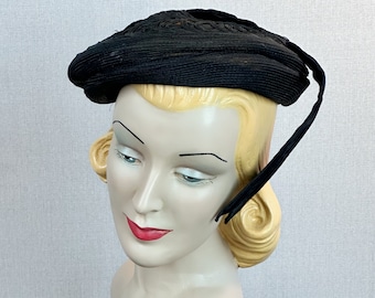 May include: A black vintage hat with a rounded crown and a wide brim. The hat is made of a woven material and features a decorative braided detail around the crown. A black ribbon extends from the hat.