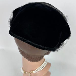 May include: A black velvet hat with a rounded crown and a small brim. The hat is trimmed with delicate black lace and embellished with clear, faceted accents. The hat is displayed on a mannequin head with a pearl necklace.
