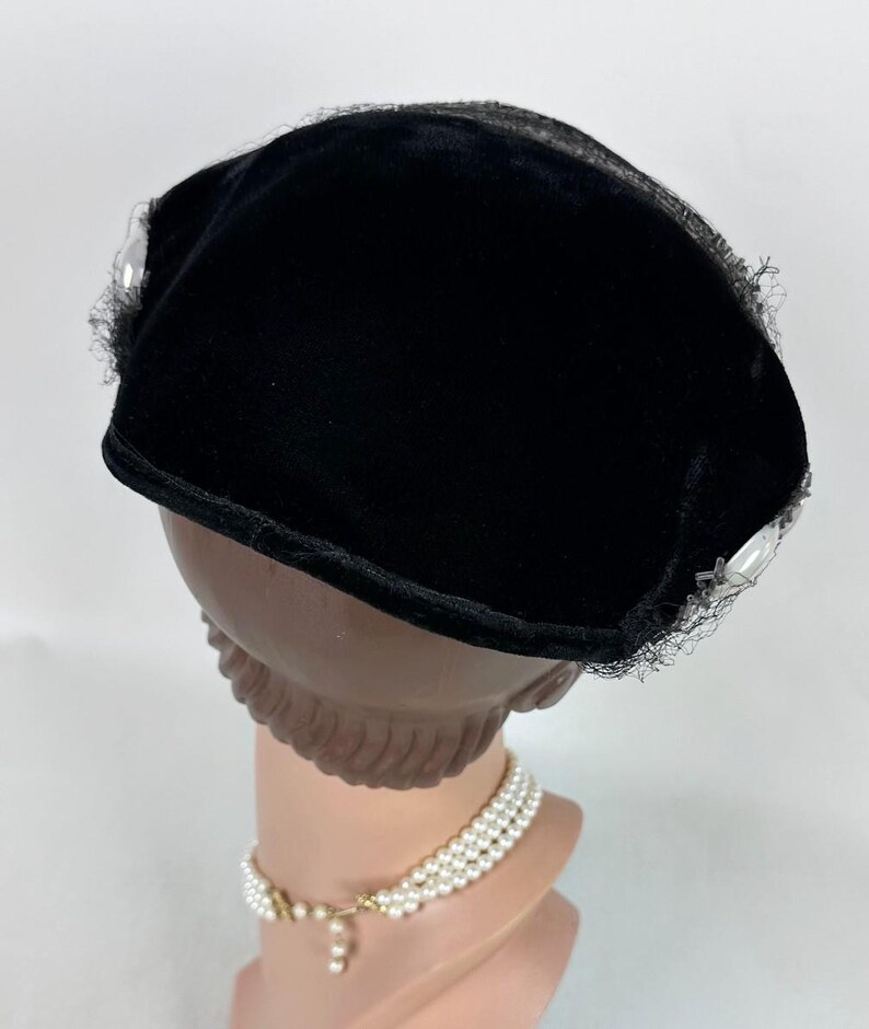 May include: A black velvet hat with a rounded crown and a small brim. The hat is trimmed with delicate black lace and embellished with clear, faceted accents. The hat is displayed on a mannequin head with a pearl necklace.