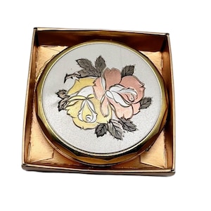 Puede incluir: Un espejo compacto vintage con un diseño floral. El compacto redondo plateado presenta un borde dorado y una tapa adornada con un motivo de rosa en tonos amarillo, rosa y cobre. Se encuentra dentro de una caja dorada.