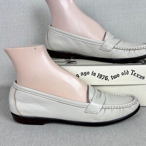 May include: Cream-coloured leather loafers with a traditional penny loafer style. The shoes feature a dark brown sole and stitched detailing. The shoes are displayed on a mannequin foot, resting on a box with text.