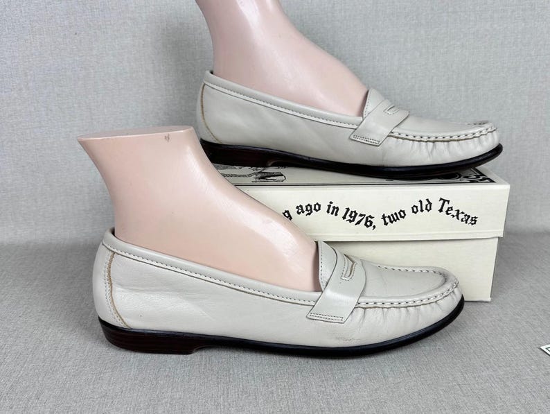 May include: Cream-coloured leather loafers with a traditional penny loafer style. The shoes feature a dark brown sole and stitched detailing. The shoes are displayed on a mannequin foot, resting on a box with text.