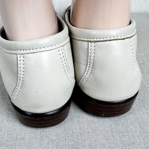 May include: A close-up view of two off-white leather loafers. The shoes feature a rounded toe, low heel, and decorative stitching. The soles are dark brown. The shoes are on a light grey surface.