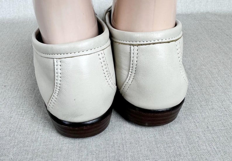 May include: A close-up view of two off-white leather loafers. The shoes feature a rounded toe, low heel, and decorative stitching. The soles are dark brown. The shoes are on a light grey surface.