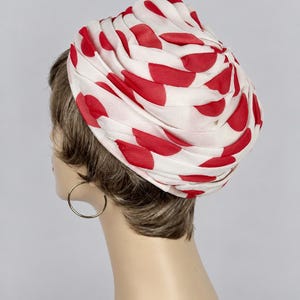 Puede incluir: Un turbante blanco con un patrón repetitivo de lunares rojos. El tocado está confeccionado con una tela ligera y semitransparente, fruncida y plisada para crear una forma redondeada y voluminosa que se asienta sobre la cabeza.