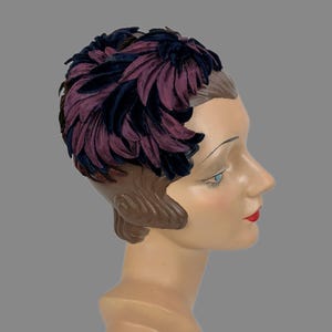 May include: A headpiece featuring layered, petal-like fabric elements in deep shades of plum purple and dark navy blue. The structured accessory covers the crown and sides of the head, displaying a dense, overlapping arrangement of dark-toned floral shapes.