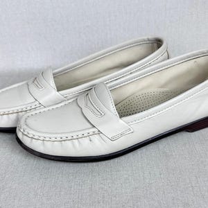 May include: A pair of white leather penny loafers. These classic shoes feature a rounded toe, a strap across the vamp, and a dark brown sole. The stitching is visible around the edges. The shoes are a timeless design.
