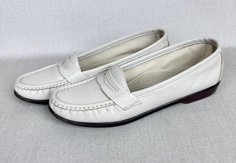 May include: A pair of white leather penny loafers. These classic shoes feature a rounded toe, a strap across the vamp, and a dark brown sole. The stitching is visible around the edges. The shoes are a timeless design.