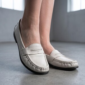 May include: Off-white leather loafers with a traditional penny-loafer design. The shoes feature a rounded toe, a strap across the vamp, and a black sole. The image shows the shoes from a low angle, highlighting their clean lines and simple elegance.