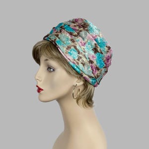May include: A turban-style hat with a pleated design. The hat features a floral pattern in shades of teal, pink, and brown. The fabric has a subtle sheen. The hat is worn by a mannequin.