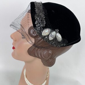 May include: Black velvet hat with a decorative beaded band and a delicate veil. The hat features three iridescent, oval-shaped embellishments. A vintage-style accessory, perfect for special occasions.