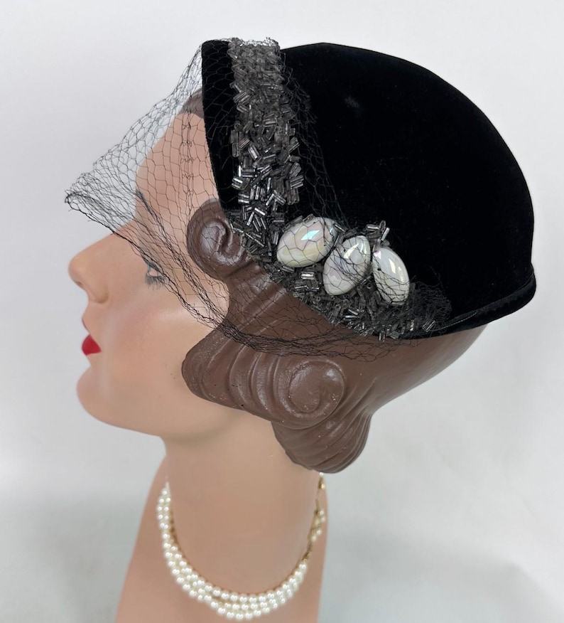 May include: Black velvet hat with a decorative beaded band and a delicate veil. The hat features three iridescent, oval-shaped embellishments. A vintage-style accessory, perfect for special occasions.