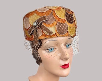 May include: Vintage hat with a circular pattern in shades of orange, brown, and yellow. A brown bow with a rhinestone accent and a delicate net veil add detail to this retro headwear.