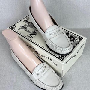 May include: A pair of white leather penny loafers with black soles are shown on a shoebox. The shoes feature a classic design with a strap detail. The shoebox has text and illustrations. The text on the box reads "shoemakers get out to play".