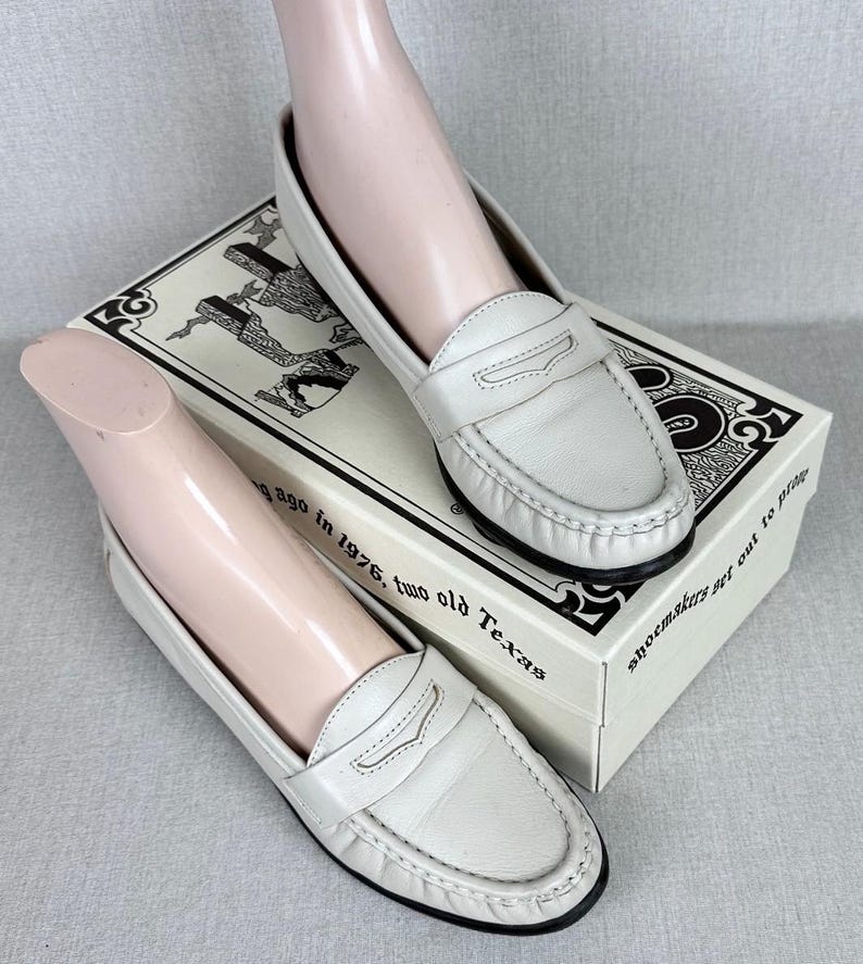 May include: A pair of white leather penny loafers with black soles are shown on a shoebox. The shoes feature a classic design with a strap detail. The shoebox has text and illustrations. The text on the box reads "shoemakers get out to play".