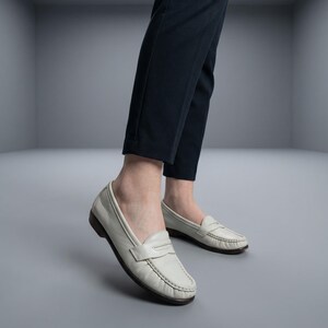 May include: White leather penny loafers with a dark brown sole. The shoes are worn with dark blue trousers. The loafers have a classic design with a strap across the top.