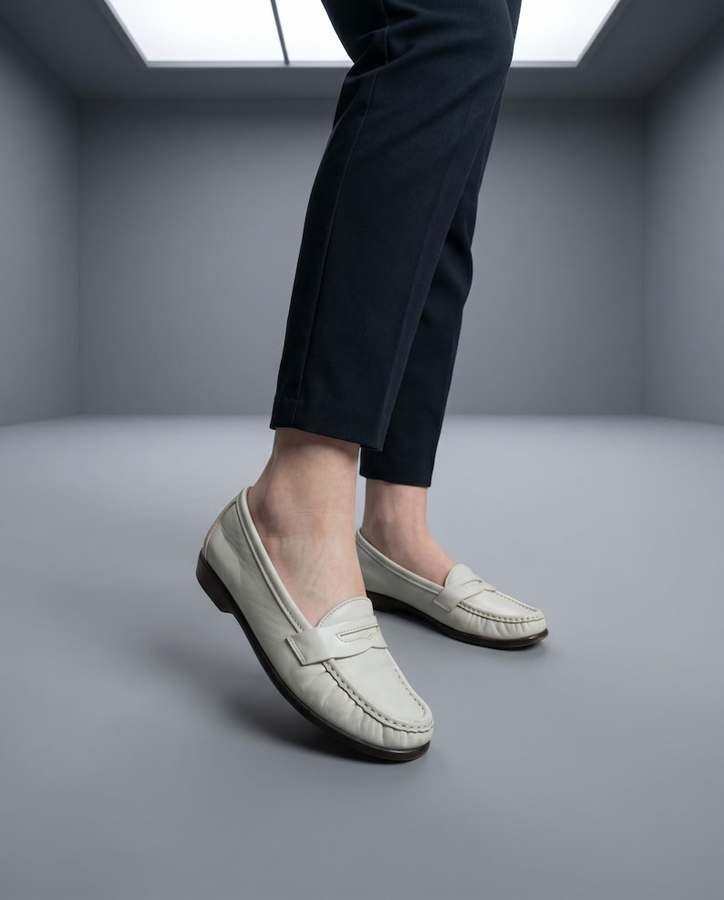 May include: White leather penny loafers with a dark brown sole. The shoes are worn with dark blue trousers. The loafers have a classic design with a strap across the top.