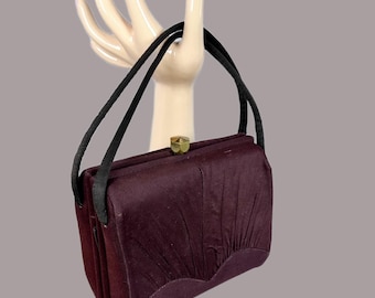 Può includere: Una borsa vintage bordeaux con manici neri e chiusura color oro. La borsa ha un design a pieghe sul davanti e una forma rettangolare strutturata. I manici sono sottili e attaccati alla parte superiore della borsa.