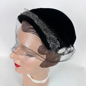 May include: A black velvet hat with a decorative beaded band and a delicate black netting veil. The hat is adorned with iridescent, oval-shaped embellishments near the brim, adding a touch of elegance to this vintage headwear.