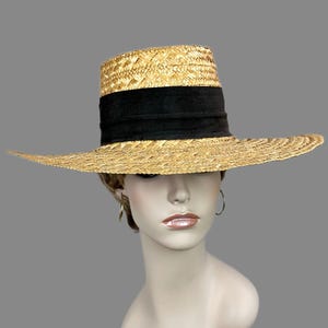 Puede incluir: Un sombrero de paja con ala ancha y una banda negra. El sombrero tiene una copa cilíndrica y un ala ancha y plana, que ofrece mucha sombra. El sombrero está hecho de paja tejida, con una banda oscura que rodea la copa.