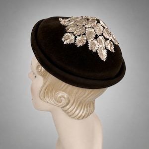 May include: A dark brown, rounded hat featuring a structured brim and a cluster of metallic silver-toned leaf-shaped embellishments arranged across the top. The hat has a smooth, felt-like texture and a deep, solid color throughout the crown.