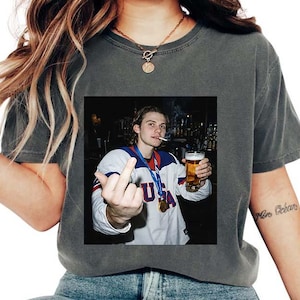 May include: Dark gray t-shirt featuring a printed image of a person holding a beer and cigarette, giving the middle finger. The person is wearing a white and blue USA jersey with a medal.