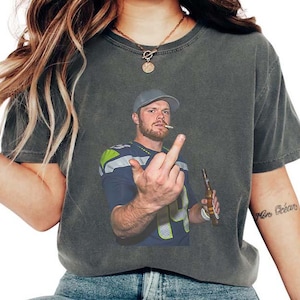 May include: Dark gray t-shirt featuring a man in a football jersey, cap, and cigarette. He is holding a beer bottle and giving the middle finger. The shirt is worn by a person with long blonde hair.