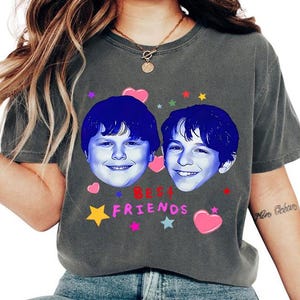 May include: Dark gray t-shirt featuring two smiling faces with blue hair, surrounded by colorful hearts, stars, and the text "BEST FRIENDS" in pink and red. A casual, comfortable tee.