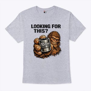 May include: A light gray t-shirt with the text "LOOKING FOR THIS?" above a cartoon illustration of a Bigfoot holding a 10mm socket. The Bigfoot is brown and smiling, with the socket in its hand.